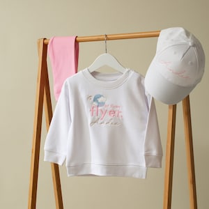 May include: White sweatshirt with a pink and blue graphic that says "First Time Flyer" and "Andie". A pink pair of leggings and a white baseball cap with the name "Andie" embroidered on it are hanging on a wooden rack.