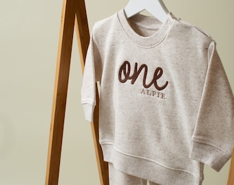 May include: A cream-colored baby onesie with long sleeves and matching pants. The onesie has the word "one" embroidered in brown on the front, with the name "Alfie" underneath.