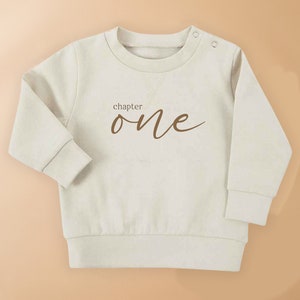May include: A cream coloured baby sweatshirt with long sleeves and a brown inscription that reads "chapter one".