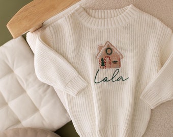 May include: Cream-colored knit sweater with a gingerbread house design embroidered on the front. The word "Lola" is embroidered in green script below the house. The sweater is displayed on a cream-colored chair with red star-shaped ornaments.