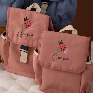 May include: Two dusty rose backpacks with ladybug and floral embroidery. The backpacks feature a flap closure, side pockets, and the name "Georgia" embroidered on the front. A blue backpack is visible in the background.