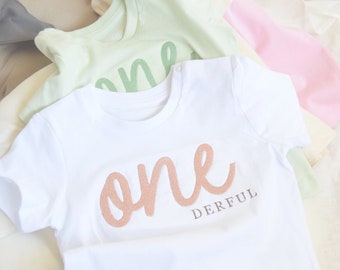 May include: White baby onesie with the word "one" in rose gold script and the word "derful" in black script below.