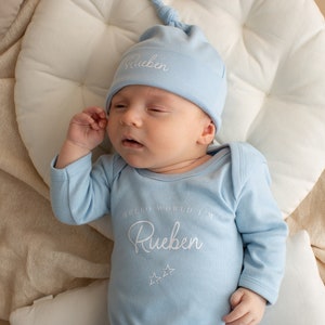 May include: A light blue baby onesie with the text "Hello World I'm Rueben" and two stars. The baby is wearing a matching light blue knotted hat with the name "Rueben" embroidered on it.