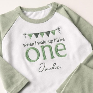 May include: A two-piece baby outfit in white and sage green. The long-sleeved top has sage green sleeves and the text "when I wake up I'll be one" with the name "Jude". The matching trousers are sage green.