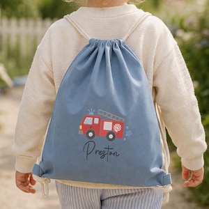 May include: A blue drawstring bag with a red fire engine design and the name "Preston". The child is wearing a cream-coloured jumper and striped trousers. The background shows a garden with a white picket fence and flowers.