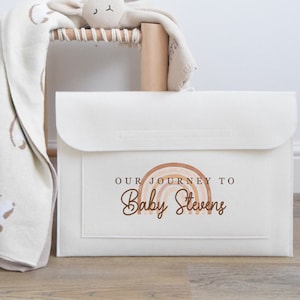 May include: Off-white fabric storage box with a flap closure. The box features a rainbow graphic and the text "OUR JOURNEY TO Baby Stevens" in brown. A blanket and stuffed animal are visible in the background.