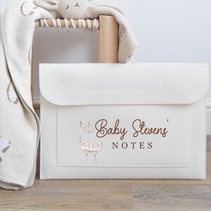 May include: Cream-colored baby notes organizer with a flap closure. The front features the text "Baby Stevens' NOTES" and a decorative illustration of a baby crib. A soft blanket and stuffed animal are visible in the background.