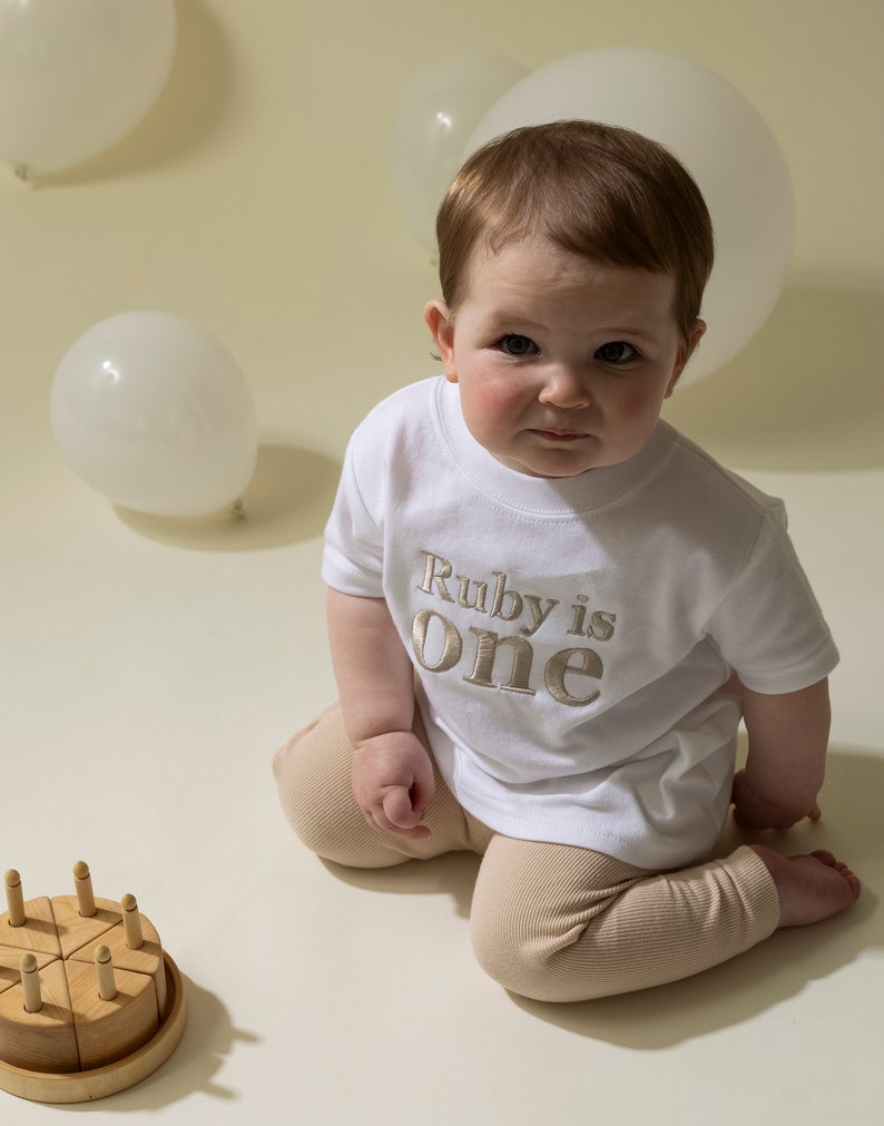 May include: A baby wearing a white t-shirt with the text "Ruby is one" in gold embroidery. The baby is sitting on a beige background with white balloons and a wooden cake.