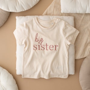 May include: A white t-shirt with the words "big sister" embroidered in pink on the front.