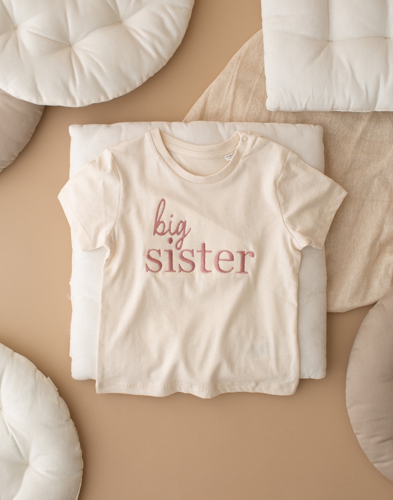 May include: A white t-shirt with the words "big sister" embroidered in pink on the front.