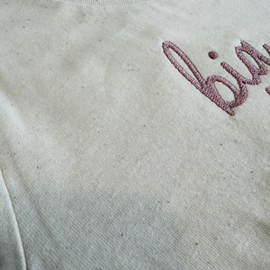 May include: A close-up of a beige t-shirt with the word "bird" embroidered in pink on the front.