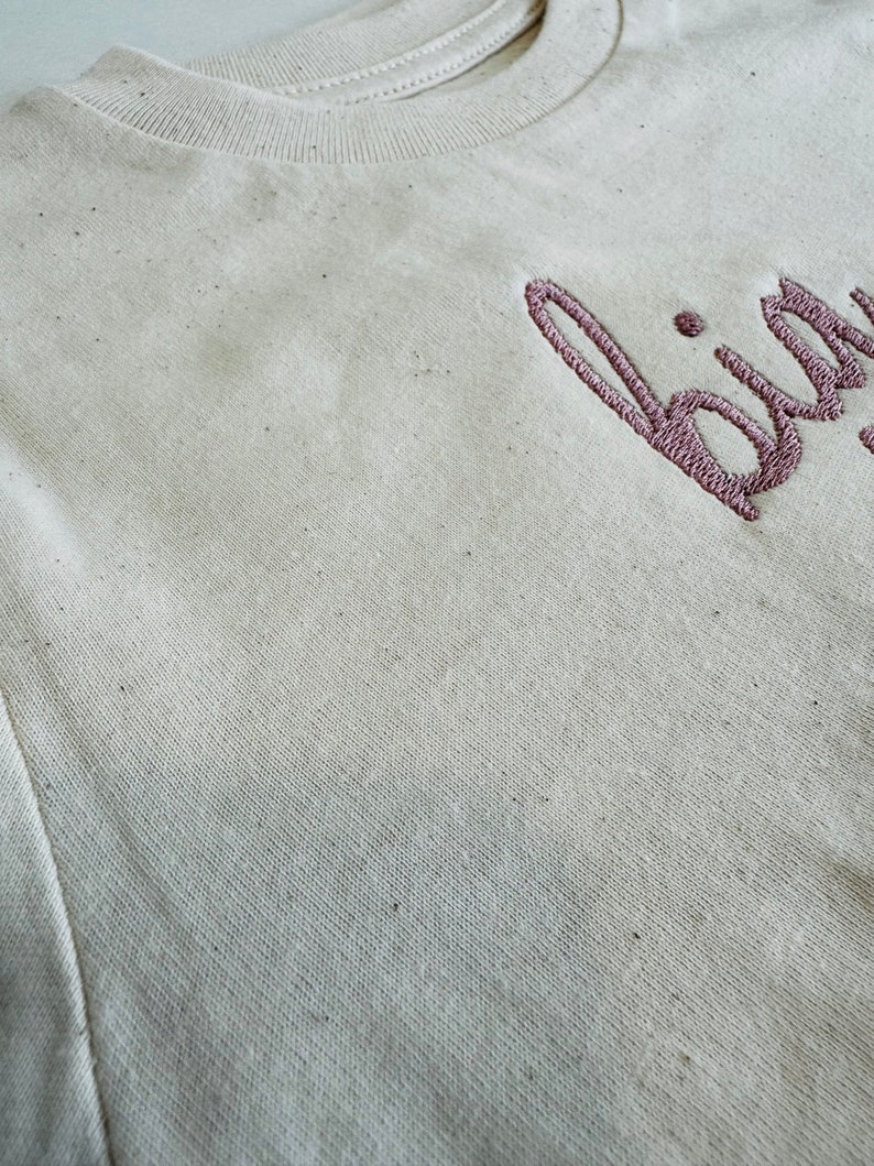 May include: A close-up of a beige t-shirt with the word "bird" embroidered in pink on the front.