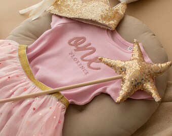 Peut inclure: Ensemble rose pour le premier anniversaire d'un bébé. Il comprend un haut à manches courtes avec le mot "one" et le nom "Florence" brodés, une jupe en tulle rose à paillettes étoiles, une couronne dorée à paillettes et une baguette magique en forme d'étoile.