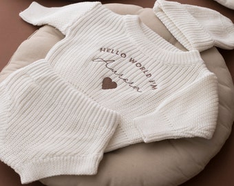 May include: White knitted baby outfit with a long-sleeved sweater, pants, and a hat. The sweater has the text "Hello World I'm Aurora" embroidered on the front with a small heart.