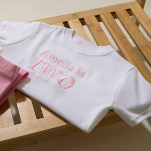 May include: White short-sleeved t-shirt with pink embroidered text that reads "Amelia is Two".