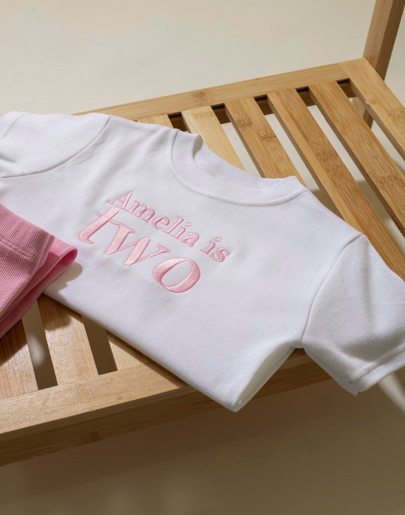 May include: White short-sleeved t-shirt with pink embroidered text that reads "Amelia is Two".