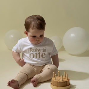 May include: A baby wearing a white t-shirt with the text "Ruby is one" in gold embroidery sits on a white surface with a wooden toy cake with candles.