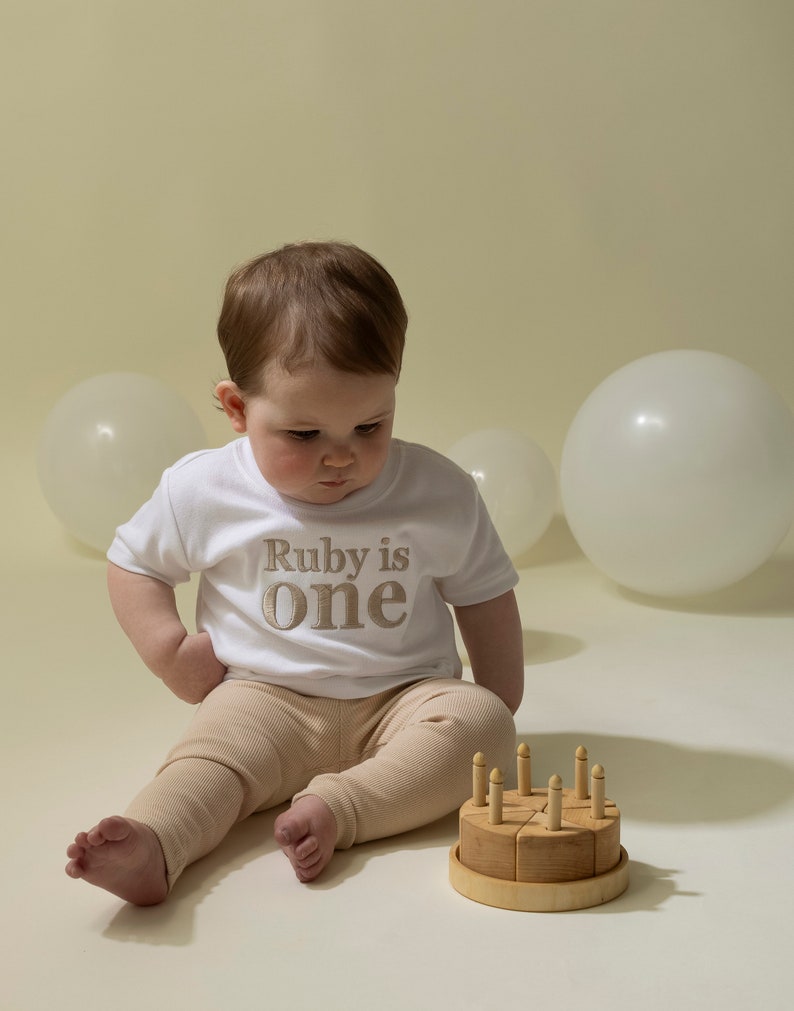 May include: A baby wearing a white t-shirt with the text "Ruby is one" in gold embroidery sits on a white surface with a wooden toy cake with candles.