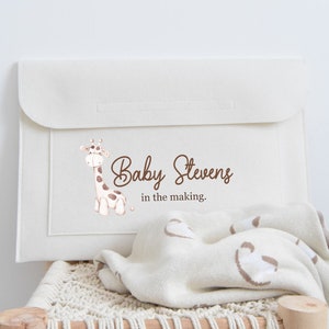 May include: Cream-colored baby changing pad cover with a giraffe illustration and the text "Baby Stevens in the making." A matching blanket with a light brown pattern is draped over a wooden chair.