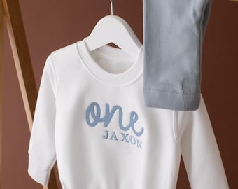 May include: White baby onesie with blue embroidered text that says "one Jaxon". The onesie is hanging on a wooden hanger with a pair of light blue leggings.