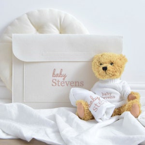 May include: A tan teddy bear wearing a white sweater and holding a white blanket, both embroidered with "baby Stevens". A white folder with the same text is next to the bear. A white, round pillow is in the background.