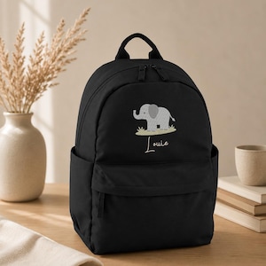 May include: A black backpack featuring an embroidered light grey elephant standing on a patch of grass. Below the elephant, the name Louie is embroidered in a white cursive script. The backpack has a top handle, two zippers, and side pockets.
