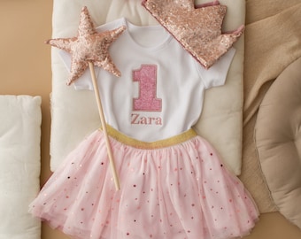 Peut inclure: Une jupe en tulle rose avec des étoiles dorées, un body blanc avec le chiffre un et le nom Zara brodé, une couronne pailletée rose et une baguette rose en forme d'étoile.