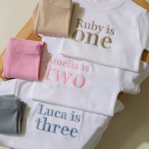 May include: Three white t-shirts with embroidered text. The top shirt says "Ruby is one" in gold thread. The middle shirt says "Amelia is two" in pink thread. The bottom shirt says "Luca is three" in blue thread.