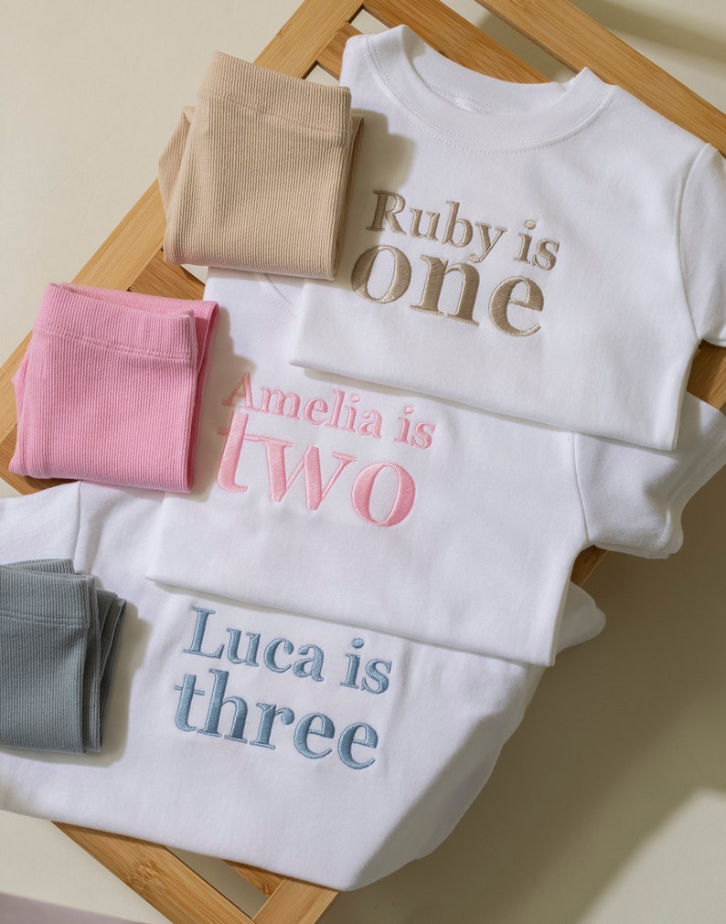 May include: Three white t-shirts with embroidered text. The top shirt says "Ruby is one" in gold thread. The middle shirt says "Amelia is two" in pink thread. The bottom shirt says "Luca is three" in blue thread.