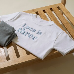 May include: White t-shirt with blue embroidered text that says "Luca is three" on a wooden surface.