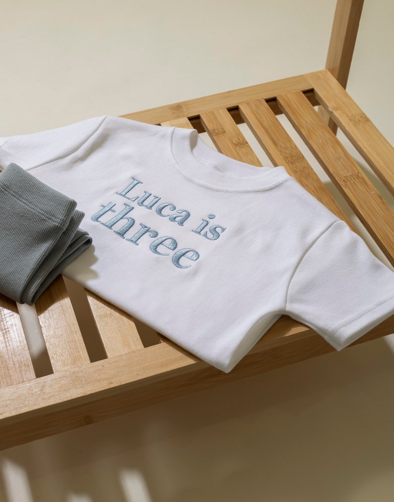 May include: White t-shirt with blue embroidered text that says "Luca is three" on a wooden surface.