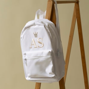 May include: White canvas backpack with a small front pocket and a zipper closure. The backpack has a crown and the initials "AS" embroidered on the front in a cream colour.