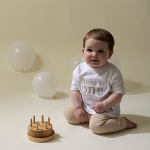 May include: A baby wearing a white shirt with the text "Ruby is one" in gold embroidery sits on a white surface with a wooden toy cake and white balloons in the background.