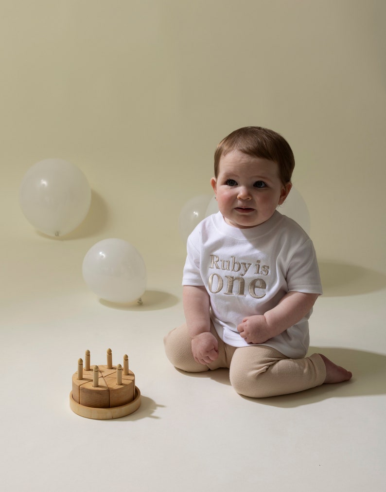 May include: A baby wearing a white shirt with the text "Ruby is one" in gold embroidery sits on a white surface with a wooden toy cake and white balloons in the background.
