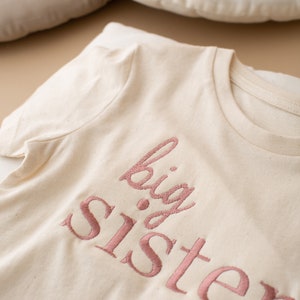 May include: A white t-shirt with the words "big sister" embroidered in pink.