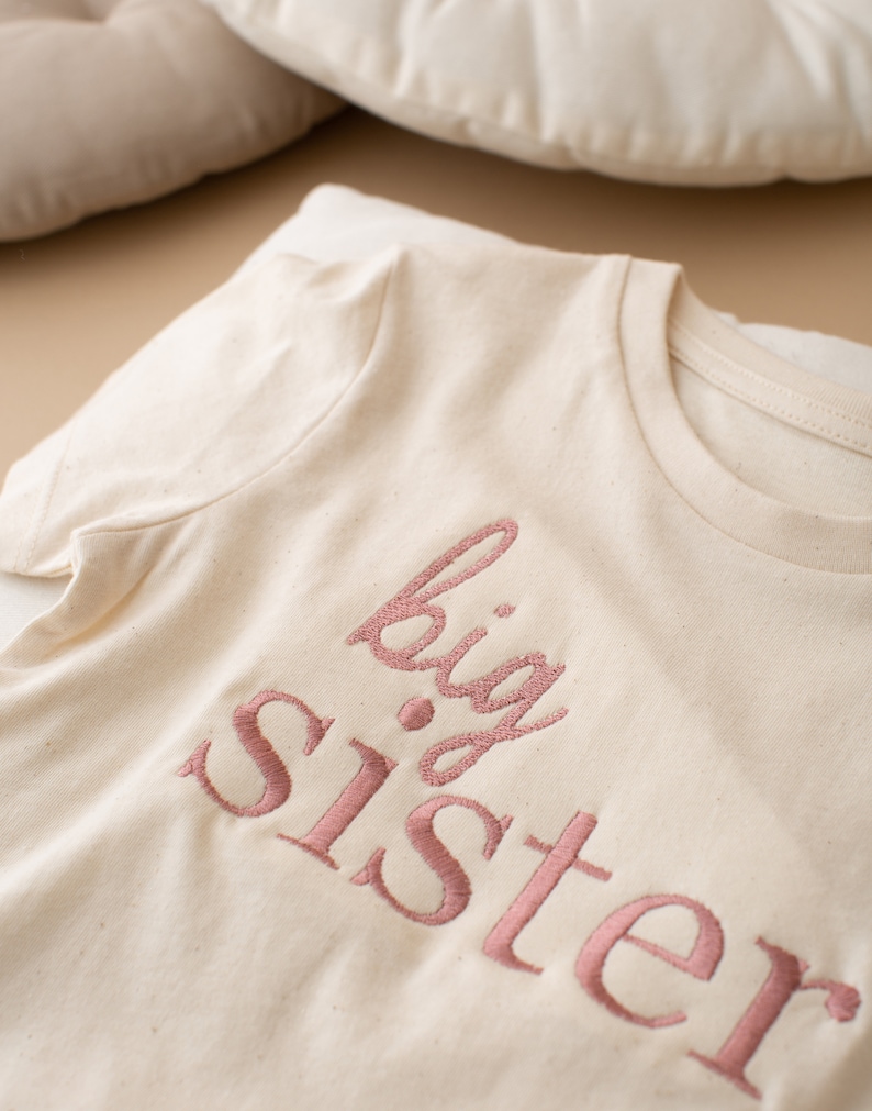 May include: A white t-shirt with the words "big sister" embroidered in pink.