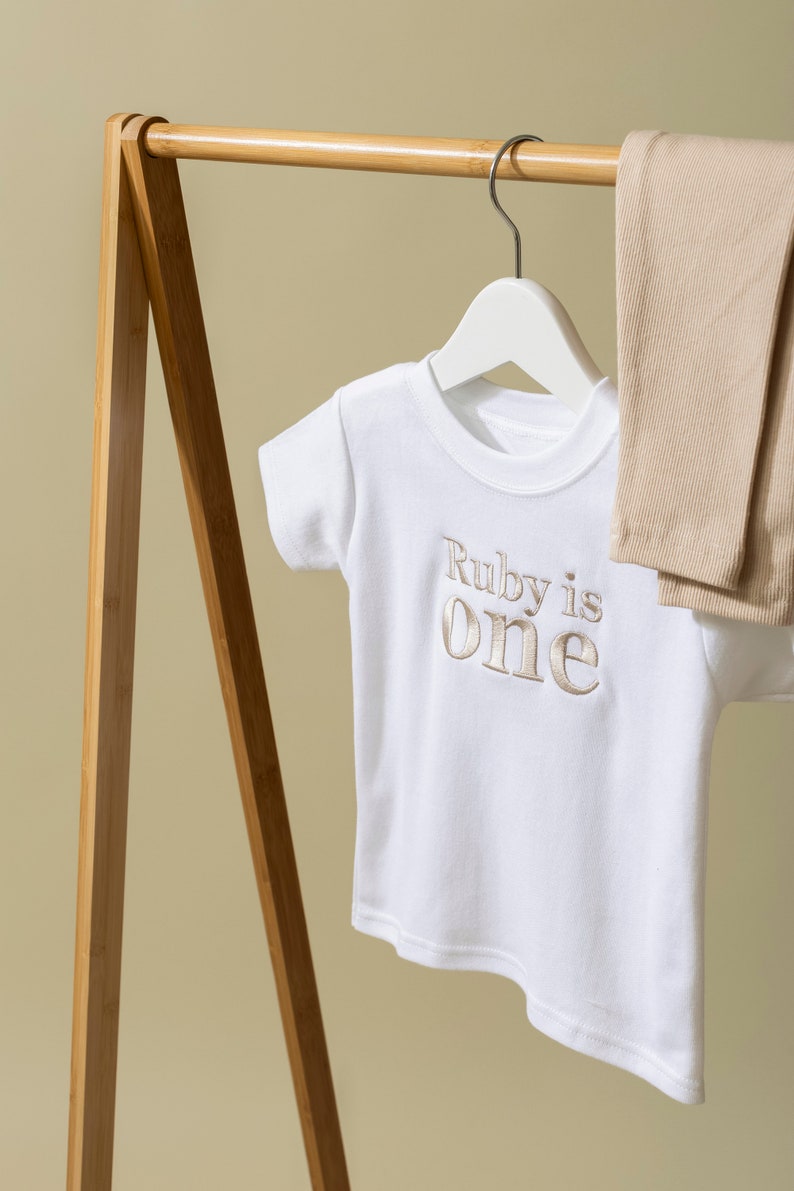 May include: White t-shirt with the text "Ruby is one" embroidered in gold on a wooden hanger. A pair of beige leggings are hanging on the hanger next to the t-shirt.