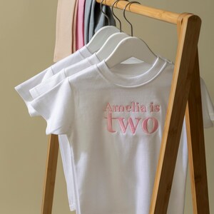 May include: White t-shirt with pink embroidered text that reads "Amelia is two" hanging on a wooden clothes rack.
