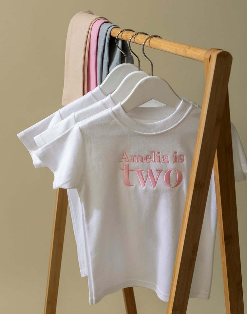 May include: White t-shirt with pink embroidered text that reads "Amelia is two" hanging on a wooden clothes rack.