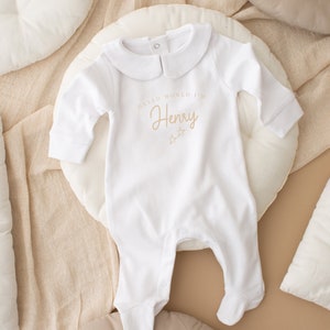 May include: White baby onesie with a peter pan collar and the text "Hello World I'm Henry" in brown lettering with two small stars.