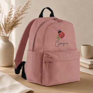 May include: A dusty rose backpack featuring a black top handle and black shoulder straps. The front displays an embroidered red ladybug with black spots resting on green leaves and pink flowers, with the name Georgia written in black script.