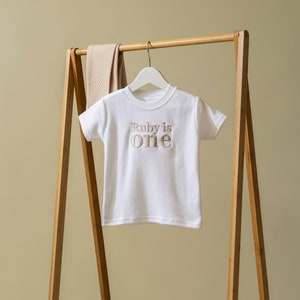 May include: White short-sleeved t-shirt with the text "Ruby is one" in brown embroidery. The shirt is hanging on a wooden clothes rack.