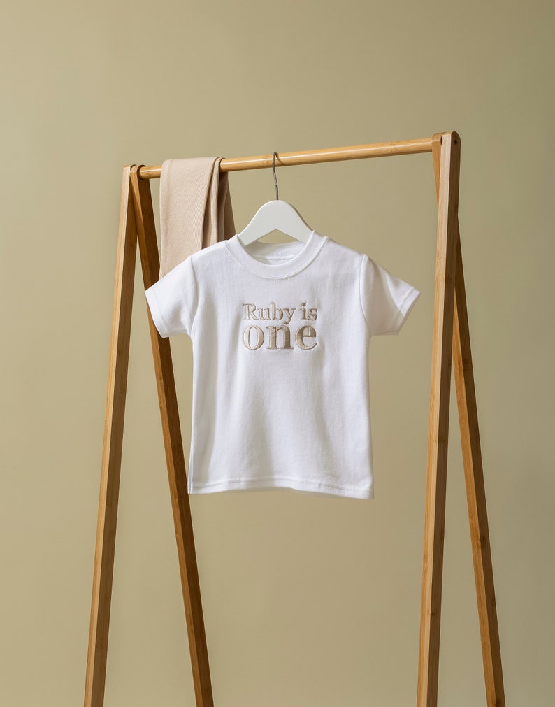 May include: White short-sleeved t-shirt with the text "Ruby is one" in brown embroidery. The shirt is hanging on a wooden clothes rack.