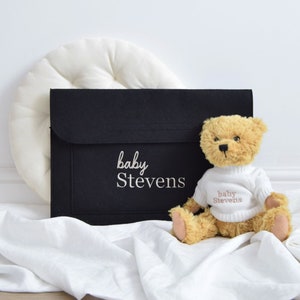 May include: A black felt baby bag with the embroidered text "baby Stevens" next to a plush teddy bear wearing a white sweater with the same text. The bear sits on a white cloth, with a white circular pillow in the background.