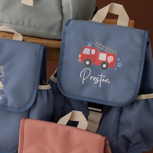 May include: Several small backpacks in shades of blue and pink are displayed. One blue backpack features a red fire truck design with the name "Preston" in white script. Other backpacks have embroidered designs, including butterflies.