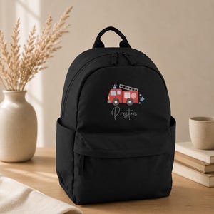 May include: A black backpack featuring a graphic of a red fire truck with a ladder and a hose, positioned above the name Preston written in a cursive script. The bag includes a top handle, a main zippered compartment, and a front pocket.