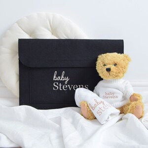 May include: A tan teddy bear wearing a white sweater and holding a white pacifier, both embroidered with "baby Stevens." A black felt case with "baby Stevens" in white script sits behind them, with a white pillow and white fabric.