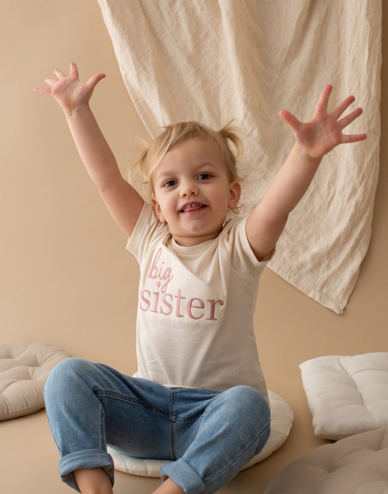 May include: A young girl wearing a white t-shirt with the words "big sister" embroidered in pink. She is wearing blue jeans and has her arms raised in the air. She is sitting on a white cushion.