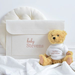 May include: A tan teddy bear wearing a white sweater with "baby Stevens" embroidered on it sits next to a white felt envelope with the same text. The items are on a white blanket, with a white, round pillow in the background.