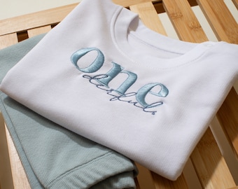 May include: White baby onesie with blue embroidered text that says "one beautiful". The onesie is folded on top of a pair of light blue pants.
